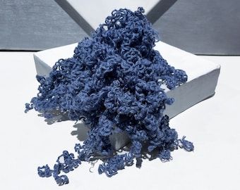 May include: A mass of blue yarn with a curly texture, cascading from a white, geometric platform. The yarn's color is a deep, muted blue, creating a contrast against the white surface. The image is well-lit, highlighting the texture of the yarn.