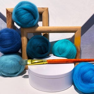 May include: An assortment of vibrant blue wool roving balls in various shades, arranged with a wooden frame and a paintbrush. The wool balls range from light turquoise to deep indigo, creating a visually appealing display for crafting projects.