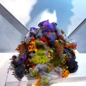 May include: A colorful, textured art piece composed of various fibers. The artwork features a mix of vibrant colors, including purple, green, orange, and blue, with small, round, yellow and orange accents. The fibers are arranged in a dense, irregular pile.