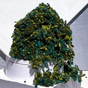 May include: A mound of textured, nubby yarn in shades of forest green, olive, and teal sits atop a smooth, white cylindrical container. The yarn has a loose, irregular, and knotted appearance with small, protruding loops throughout the pile.