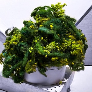 May include: A vibrant arrangement of green and yellow wool fibers, resembling a plant, fills a white ceramic pot. The wool is a mix of textures and shades, creating a visually rich display. The pot sits on a white, textured platform.