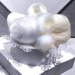 May include: A decorative arrangement of white and iridescent fiber, resembling snow, atop a small, round, silver container. A clear, snowflake-shaped ornament sits on the container's edge, casting shadows. The composition is set against a white and silver backdrop.