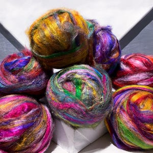 May include: A collection of colorful yarn balls in shades of yellow, green, purple, pink, and red. The yarn appears to be a blend of fibers, with a soft, textured appearance. The balls are arranged on a white surface.