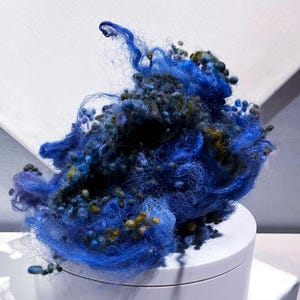 May include: A vibrant, abstract composition of blue and black fibers with yellow and green accents. The textured material is arranged in a cloud-like form, set against a white background and a white cylindrical base.