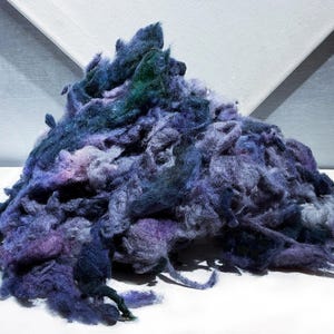 May include: A pile of dyed fiber in shades of blue, purple, and green. The fibers are loosely arranged, creating a textured, cloud-like appearance. The fibers are on a white surface.