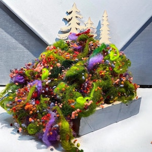 May include: A colorful, textured art piece featuring a mound of green, purple, orange, and red fibers. Wooden tree cutouts are placed on top of the fiber mound, which sits in a small, gray rectangular container. The background is a light gray.