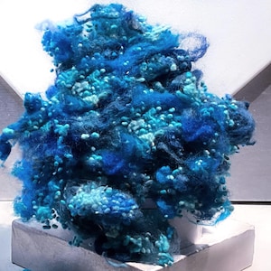 May include: A close-up of a textured, abstract sculpture composed of blue and turquoise fibers. The fibers are densely packed with small, white, bead-like elements, creating a visually rich and tactile surface. The sculpture sits on a white, geometric base.