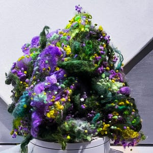 Può includere: Un vibrante mucchio di filato colorato nelle tonalità del viola, verde e giallo. Il filato strutturato è disposto a cumulo, con una varietà di trame e piccole decorazioni rotonde. Il filato è su una superficie bianca.