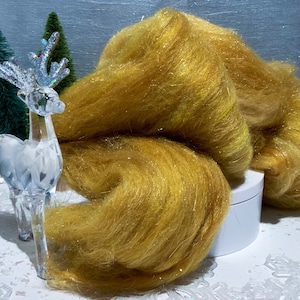 May include: A pile of golden-yellow, sparkly fiber, possibly yarn or roving, sits on a white surface. A clear, crystal-like reindeer figurine and two small green Christmas trees are in the background, suggesting a holiday theme.