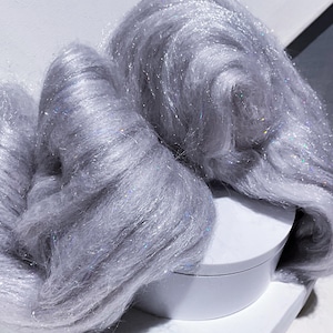 May include: Close-up of two large, fluffy bundles of silver-gray yarn with iridescent glitter strands. The yarn is arranged on a white, round surface, creating a soft, textured appearance. Ideal for fiber art projects.