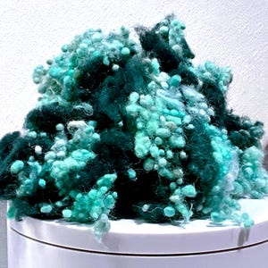 May include: A mound of teal and dark green yarn with a textured, knobbly appearance. The yarn is arranged on a white surface, with a white background visible behind it. The yarn appears to be a blend of colours, creating a visually interesting texture.
