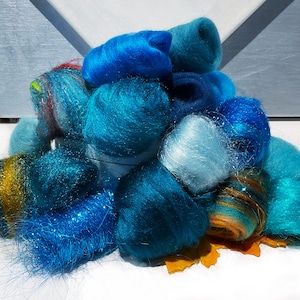 May include: A collection of blue and teal fiber bundles, varying in shades and textures, including some with glitter accents. The fibers are arranged in a pile, with a few yellow-orange leaves at the base. The background is a light blue and white.