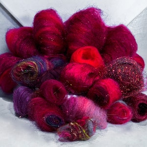 May include: A collection of colorful, fluffy yarn in shades of pink, red, and purple. The yarn is arranged in a loose pile, with some strands of glitter yarn visible.