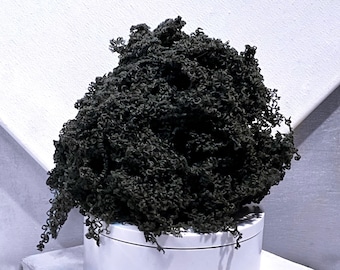 May include: A dark green, textured, cloud-like object, possibly yarn or fabric, sits atop a white cylindrical base. The material has a dense, curly appearance, with a slightly irregular shape. The background is a white surface and a gray wall.