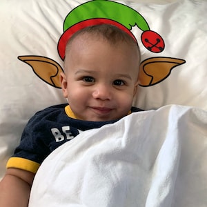 May include: A smiling baby lies on a white pillow with a Christmas elf hat design. The hat is green and red with a gold bell. The baby wears a navy blue shirt with yellow trim and the word "BE" visible.