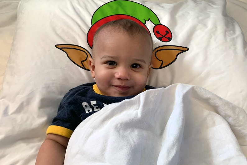 May include: A smiling baby lies on a white pillow with a Christmas elf hat design. The hat is green and red with a gold bell. The baby wears a navy blue shirt with yellow trim and the word "BE" visible.