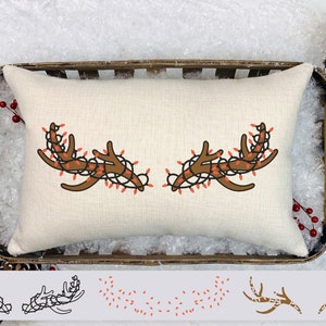 May include: A rectangular, off-white pillow with a brown antler design wrapped in red Christmas lights. The pillow is in a wooden tray with pinecones and faux snow, suggesting a holiday theme.