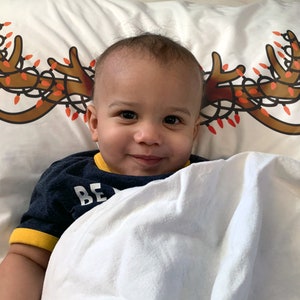 May include: A smiling child is lying on a white pillow with a Christmas antler design. The antlers are brown with red and orange lights. The child is wearing a navy blue shirt with yellow trim and the word "BE" visible.