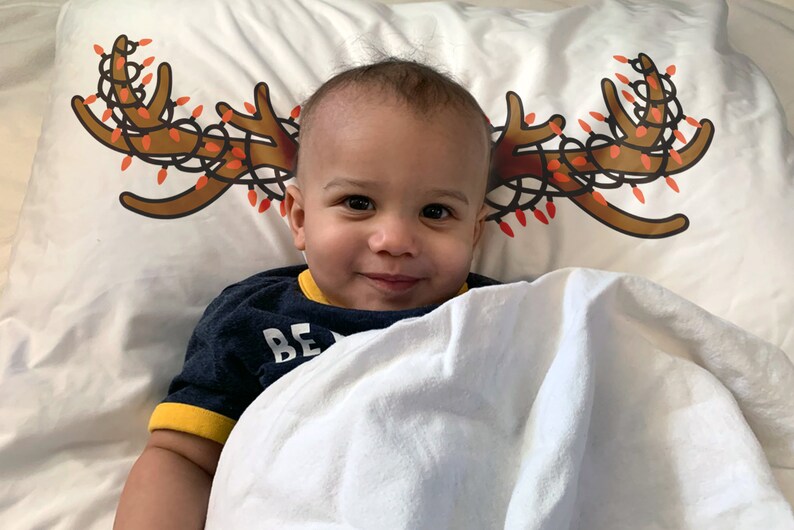 May include: A smiling child is lying on a white pillow with a Christmas antler design. The antlers are brown with red and orange lights. The child is wearing a navy blue shirt with yellow trim and the word "BE" visible.