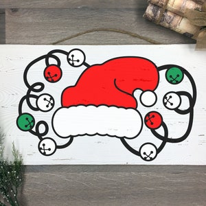 May include: A rectangular wooden sign featuring a red Santa hat with a white brim and black bells with red and green accents. The sign is white with a distressed finish and a jute rope for hanging.