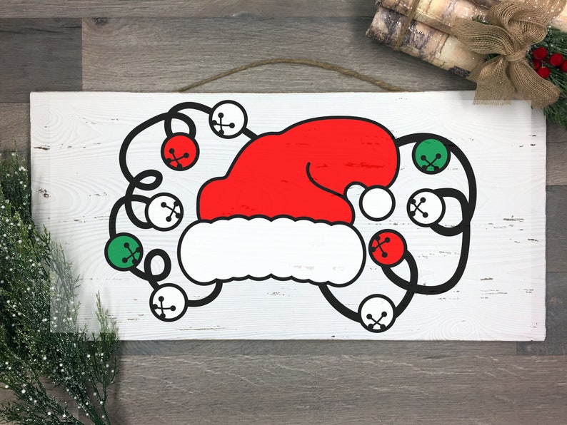 May include: A rectangular wooden sign featuring a red Santa hat with a white brim and black bells with red and green accents. The sign is white with a distressed finish and a jute rope for hanging.