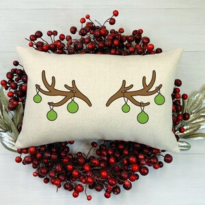 May include: A rectangular pillow with a cream-colored background features a brown antler design with green ornaments. The pillow is surrounded by a wreath of red berries and silver leaves, creating a festive holiday theme.