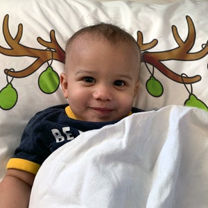 May include: A smiling baby lies on a white pillow decorated with a brown antler design and green Christmas ornaments. The baby wears a navy blue shirt with yellow trim and the word "BE" visible.