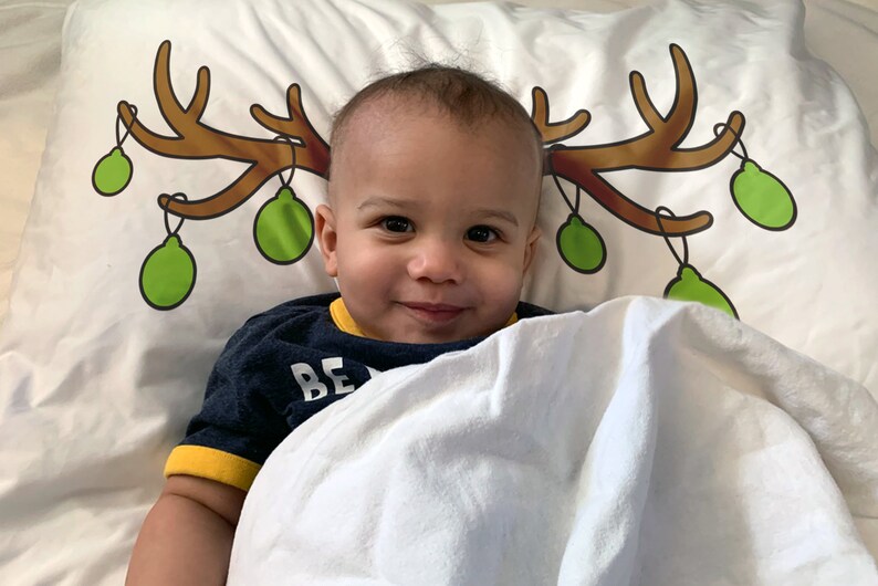 May include: A smiling baby lies on a white pillow decorated with a brown antler design and green Christmas ornaments. The baby wears a navy blue shirt with yellow trim and the word "BE" visible.