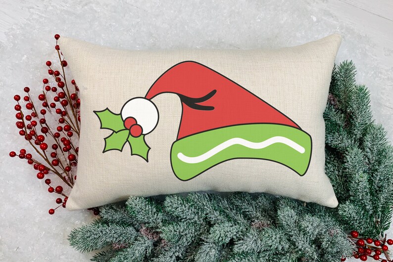 May include: A rectangular decorative pillow with a Christmas elf hat design. The hat is red with a green brim and a white ball. Holly with red berries is on the left side of the hat. The pillow is on a light background with greenery.