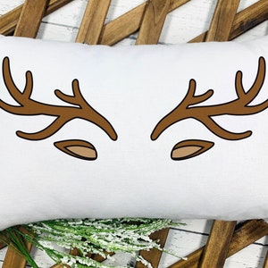 May include: White rectangular pillow with a brown antler and ear design. The antlers are stylized and symmetrical, with a simple, cartoonish aesthetic. The pillow is placed on a wooden background with floral accents.