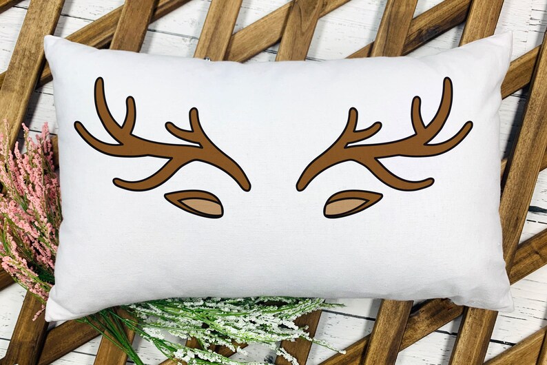 May include: White rectangular pillow with a brown antler and ear design. The antlers are stylized and symmetrical, with a simple, cartoonish aesthetic. The pillow is placed on a wooden background with floral accents.