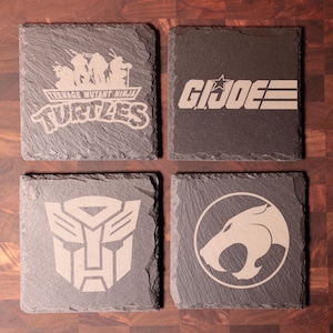 May include: Four square slate coasters with etched designs. The designs include the Teenage Mutant Ninja Turtles logo, the G.I. Joe logo, a Transformers symbol, and a Thundercats emblem. The coasters are dark gray.