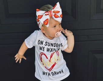 May include: A baby girl wearing a white t-shirt with the text "On Sundays We Watch Chiefs With Daddy" and a red heart with the word "Chiefs" inside. She is wearing a red and white bow with the word "Chiefs" printed on it. She is wearing red sneakers and black leggings.