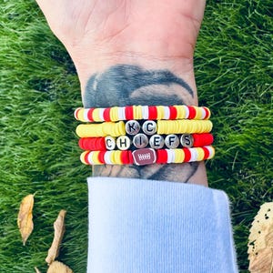 Könnte beinhalten: Ein Stapel aus vier Armbändern mit roten, gelben und weißen Perlen. Die Armbänder buchstabieren "KC CHIEFS" mit einem Fußball-Anhänger.
