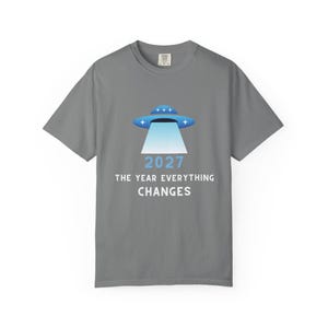 Könnte beinhalten: Graues T-Shirt mit einem blau-weißen UFO-Design und dem Text "2027 THE YEAR EVERYTHING CHANGES". Das T-Shirt hat einen Rundhalsausschnitt und kurze Ärmel. Die Grafik ist mittig auf der Vorderseite.