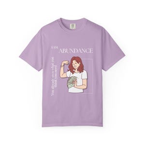 May include: A lilac-colored t-shirt with the words "I AM ABUNDANCE" and an illustration of a person flexing their arm while holding money. The text "You already own what you think you need" is also printed on the shirt.