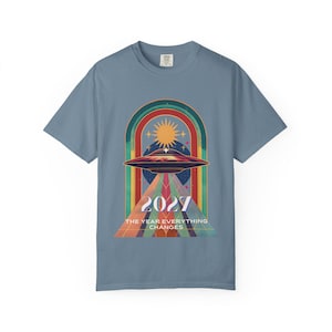 Puede incluir: Camiseta azul con un diseño retro. Presenta un OVNI, un sol y un arcoíris. El gráfico incluye el año "2027" y el texto "THE YEAR EVERYTHING CHANGES" en un estilo vintage colorido.