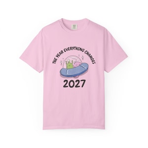 May include: A light pink t-shirt featuring a cartoon graphic of a blue UFO with two aliens inside. The text "THE YEAR EVERYTHING CHANGES" arches above the UFO, with "2027" printed below in black.