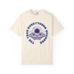 May include: Cream-colored t-shirt featuring a circular graphic with an astronaut in a spaceship. The text "THE YEAR EVERYTHING CHANGES 2027" is printed around the spaceship in blue. The shirt has short sleeves and a crew neck.