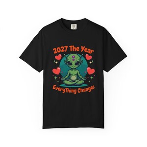 May include: Black t-shirt featuring a cartoon alien meditating with the text "2027 The Year Everything Changes". The alien is green with purple spots, surrounded by red hearts and stars. The design is in a circular frame.