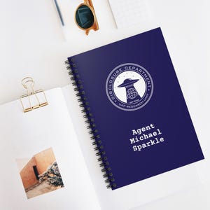 Może przedstawiać: Niebieski notatnik w oprawie spiralnej z napisem "DISCLOSURE DEPARTMENT UAP RESEARCH UNIT" w okrągłym wzorze. Notatnik zawiera również imię i nazwisko "Agent Michael Sparkle" w białym tekście.