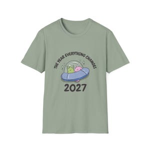 May include: Sage green t-shirt featuring a cartoon illustration of a UFO with two aliens inside. The text "THE YEAR EVERYTHING CHANGES" arcs above the UFO, with the year "2027" below.