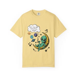 May include: Yellow t-shirt featuring a cartoon alien smoking a cigar while seated on a crescent moon. The design includes planets, stars, and the text "2027 The year Everything changes" in a thought bubble.
