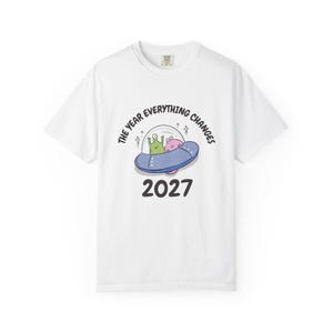 May include: White t-shirt featuring a cartoon illustration of a blue UFO with two aliens inside. The text "THE YEAR EVERYTHING CHANGES" arches above the UFO, with "2027" below. The shirt has a crew neck.