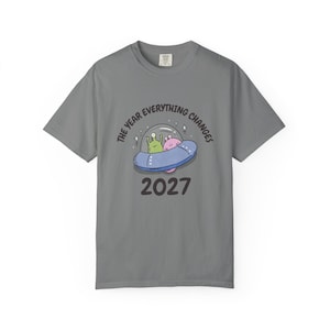 May include: Gray t-shirt with a graphic of a UFO carrying two aliens. The text "THE YEAR EVERYTHING CHANGES" arches above the UFO, and "2027" is printed below. The shirt is made of a soft material.