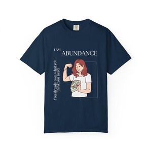 May include: Navy blue t-shirt with the text "I AM ABUNDANCE" and a graphic of a person flexing their arm while holding money. The text "You already own what you think you need" is printed vertically on the left side.
