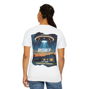Könnte beinhalten: Weißes T-Shirt mit einer Grafik eines UFOs, das einen Lichtstrahl auf eine Vorstadtszene richtet. Die Grafik enthält das Jahr "2027" und den Text "The Year Everything Changes".