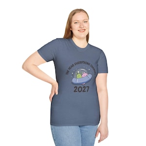May include: A blue t-shirt featuring a cartoon graphic of a UFO with two aliens inside. The text "THE YEAR EVERYTHING CHANGES" is above the UFO, and the year "2027" is below. The shirt is a solid color.