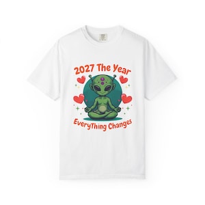 May include: White t-shirt featuring a green alien meditating with the text "2027 The Year Everything Changes" in red and yellow. The design includes red hearts and green stars. The shirt is a crew neck.