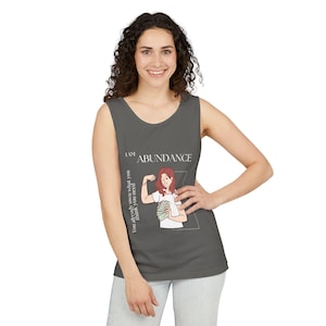 Könnte beinhalten: Graues Tanktop mit der Aufschrift "I AM ABUNDANCE" und einer Grafik einer Frau, die ihren Arm beugt und Geld hält. Der Text "You already own what you think you need" ist vertikal auf der linken Seite aufgedruckt.